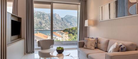 Un salon moderne avec un canapé, des fauteuils, une petite table sur laquelle on a disposé des fruits et un téléviseur. Une grande fenêtre offre une vue sur les montagnes et un lac.