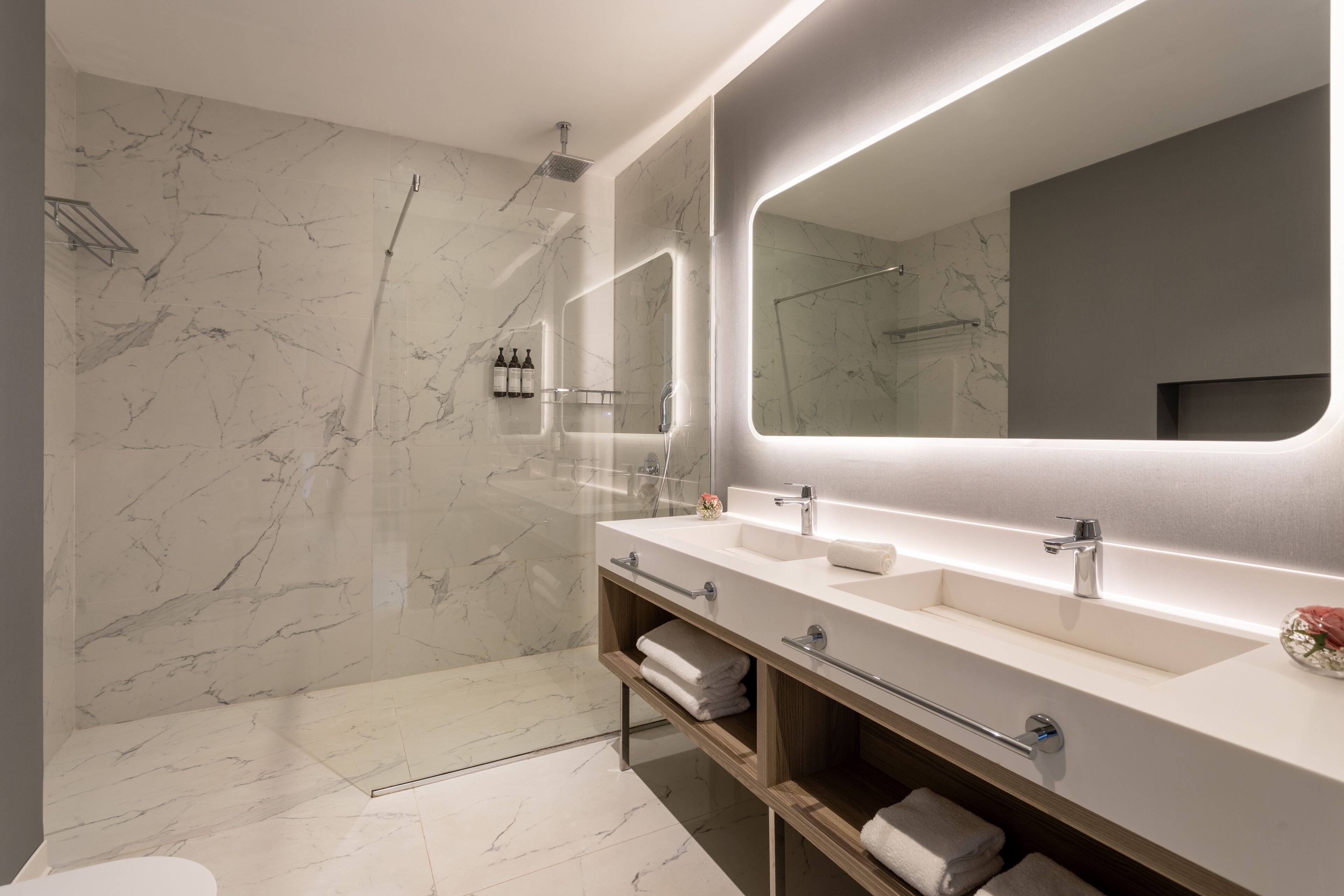 A modern bathroom with marble walls, a large mirror, dual sinks, and a shower area.