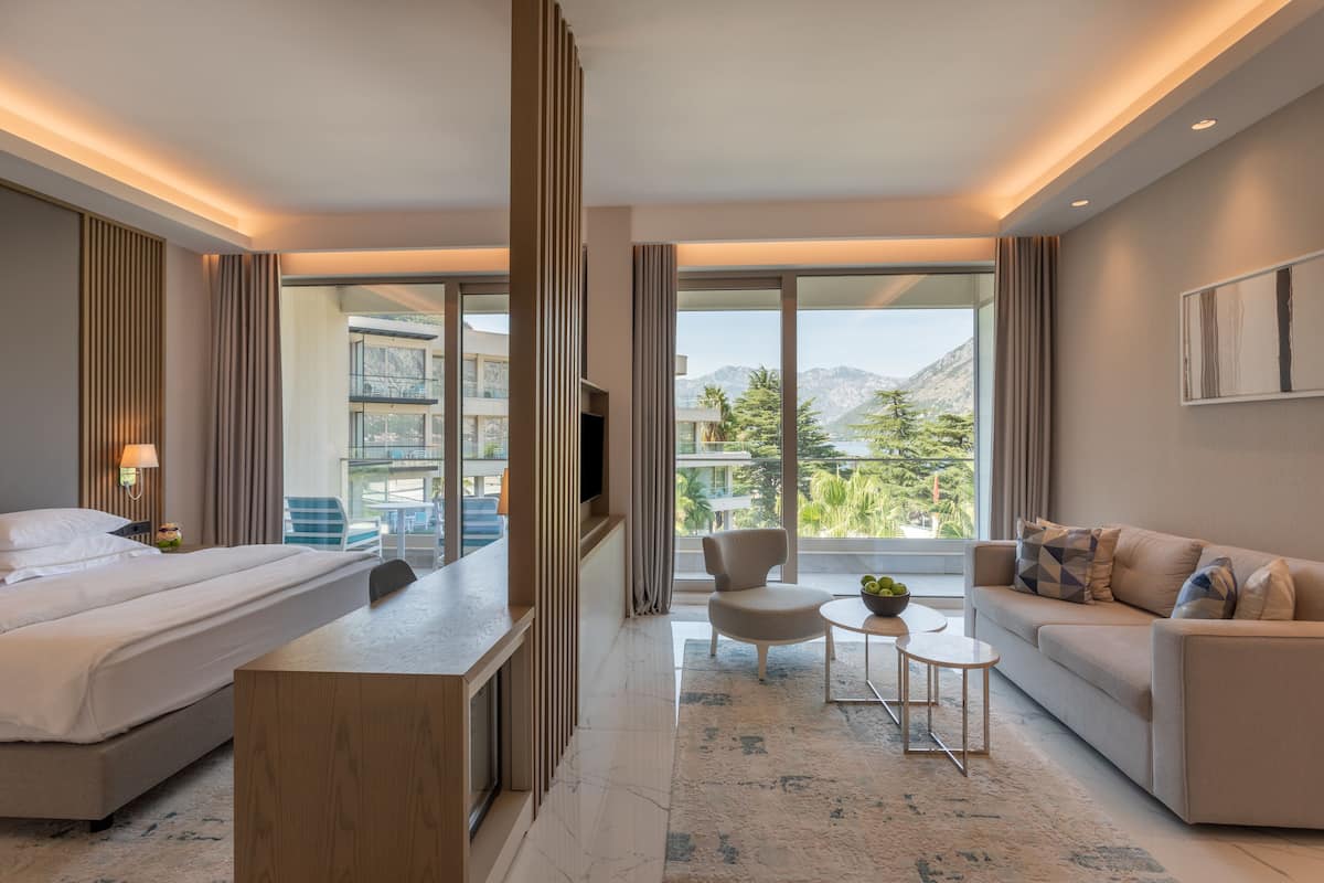 Une chambre d’hôtel moderne équipée d’un lit, d’un bureau, d’un canapé et d’une télévision. La chambre dispose de grandes fenêtres offrant une vue sur les montagnes.
