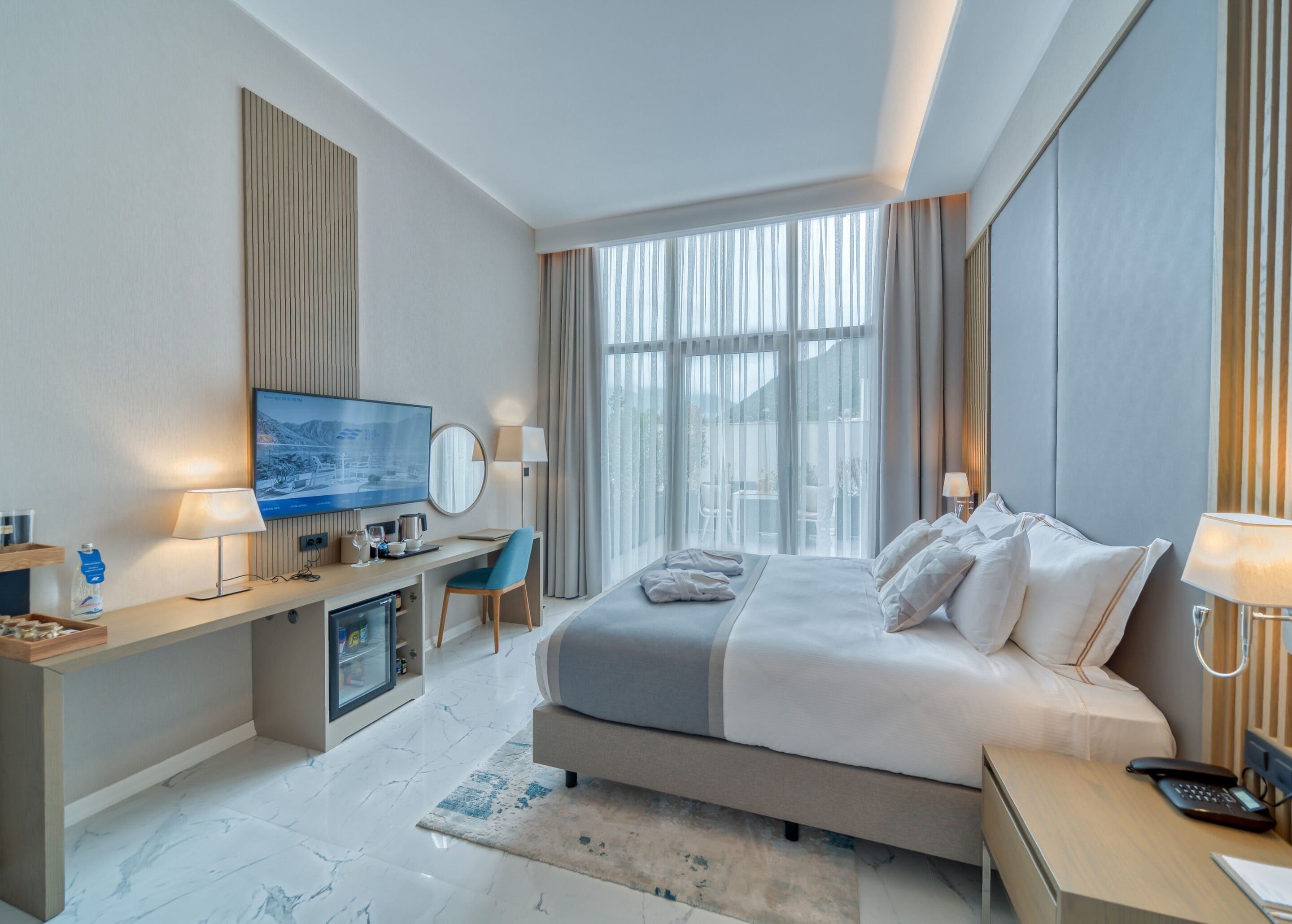 A modern hotel room with a large bed, a desk with a TV, a seating area, and a view through large windows.