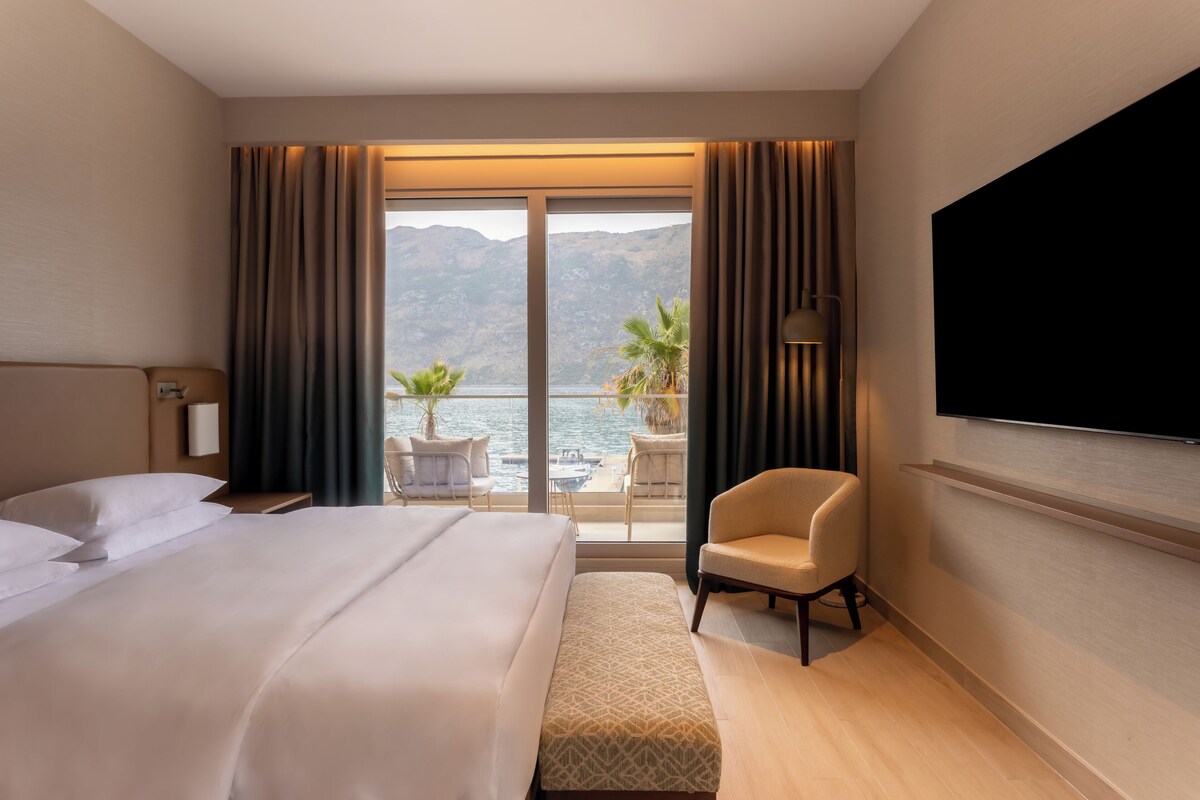 Habitación de hotel con una cama grande, un televisor en la pared, una silla y un balcón con vistas a las montañas y al agua.