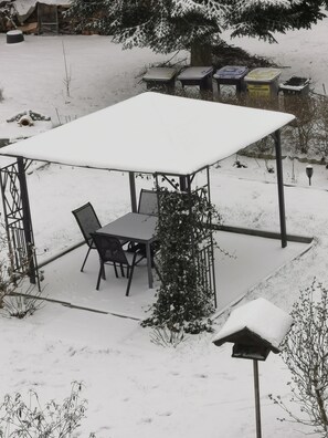 A covered outdoor seating area with a table and chairs, surrounded by snow-covered ground and trees. - Apartment Wiesenblick - Apartment (Schenkendöbern)