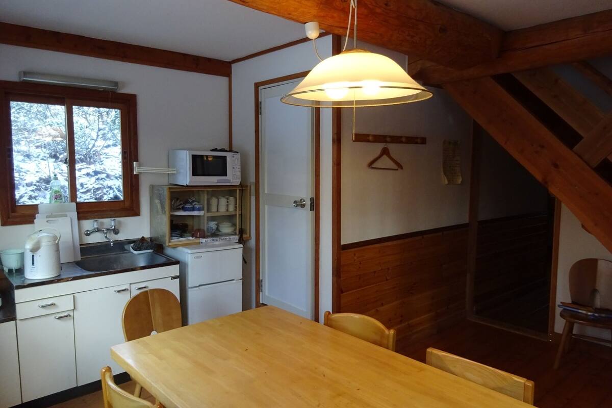 Cocina compacta con una mesa de madera, sillas, un microondas y una lámpara colgante.