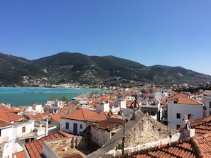 A coastal town with terracotta-roofed buildings, a clear blue sea, and a mountainous backdrop.