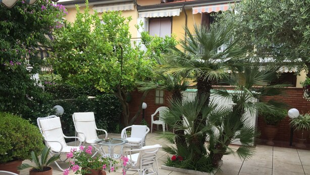 An outdoor patio with white wicker furniture, a glass table, and various potted plants. - Casa Matilde (Massa)
