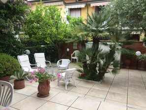 Un patio al aire libre con mobiliario de mimbre blanco, una mesa de vidrio y diversas plantas en macetas.