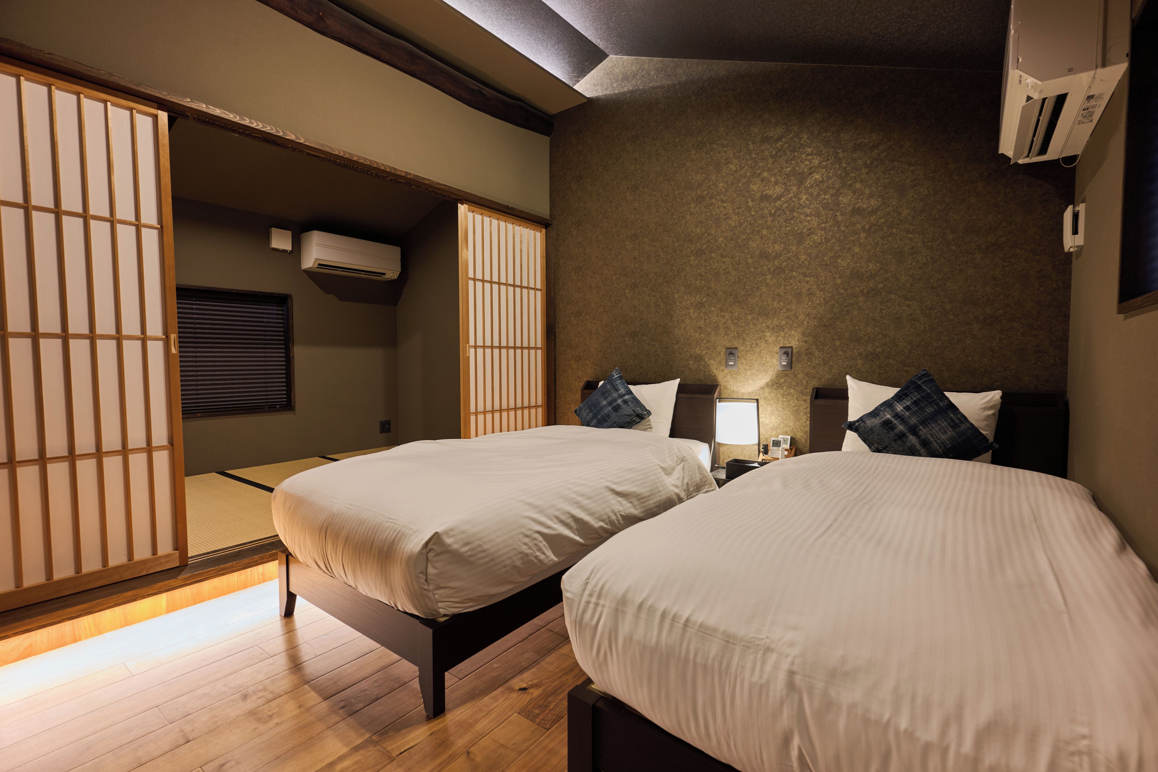 A hotel room with two beds, a wooden floor, a sliding shoji screen, and a wall-mounted air conditioner.