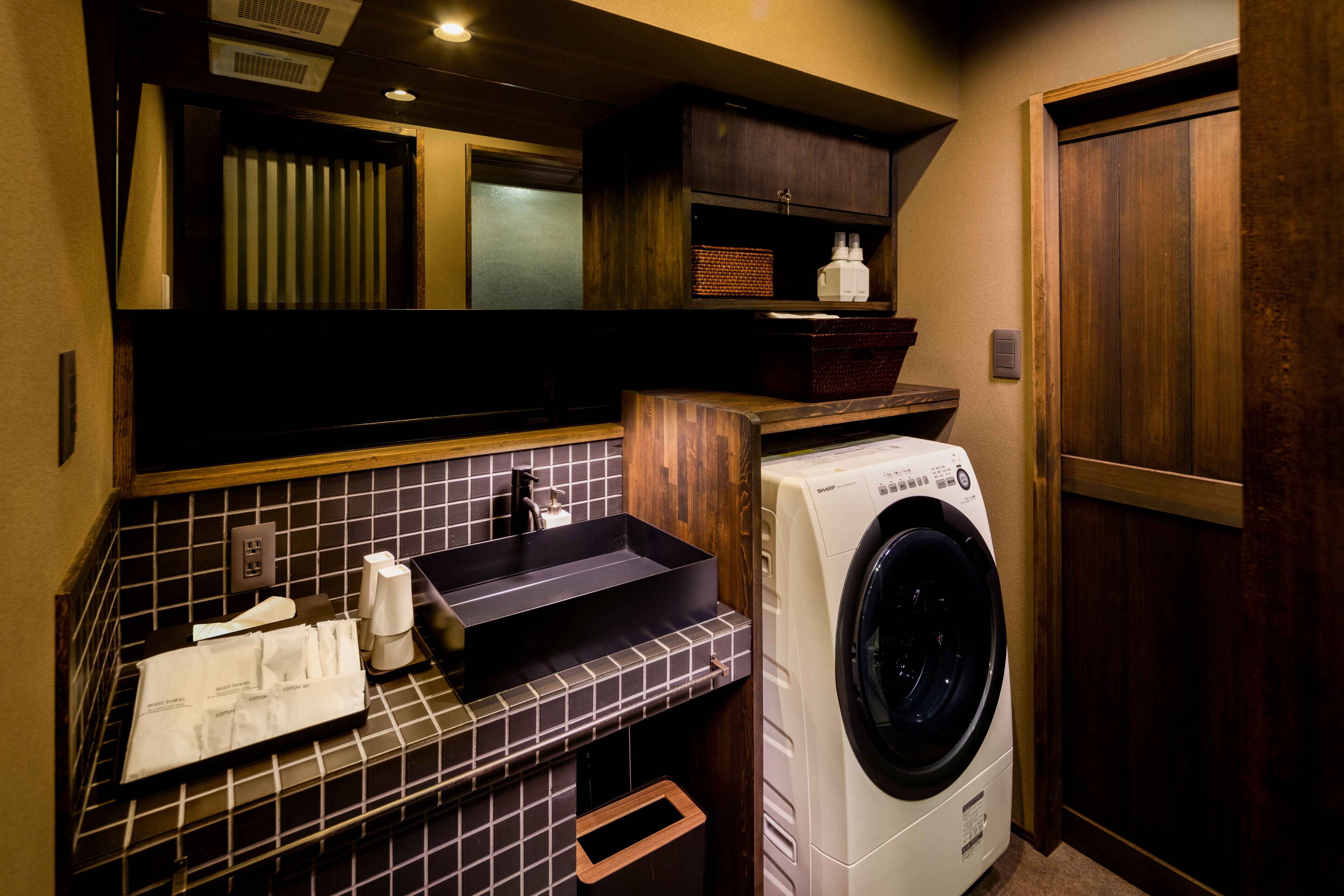A compact hotel bathroom with a washing machine, a sink with a black basin, and a mirror above the sink.