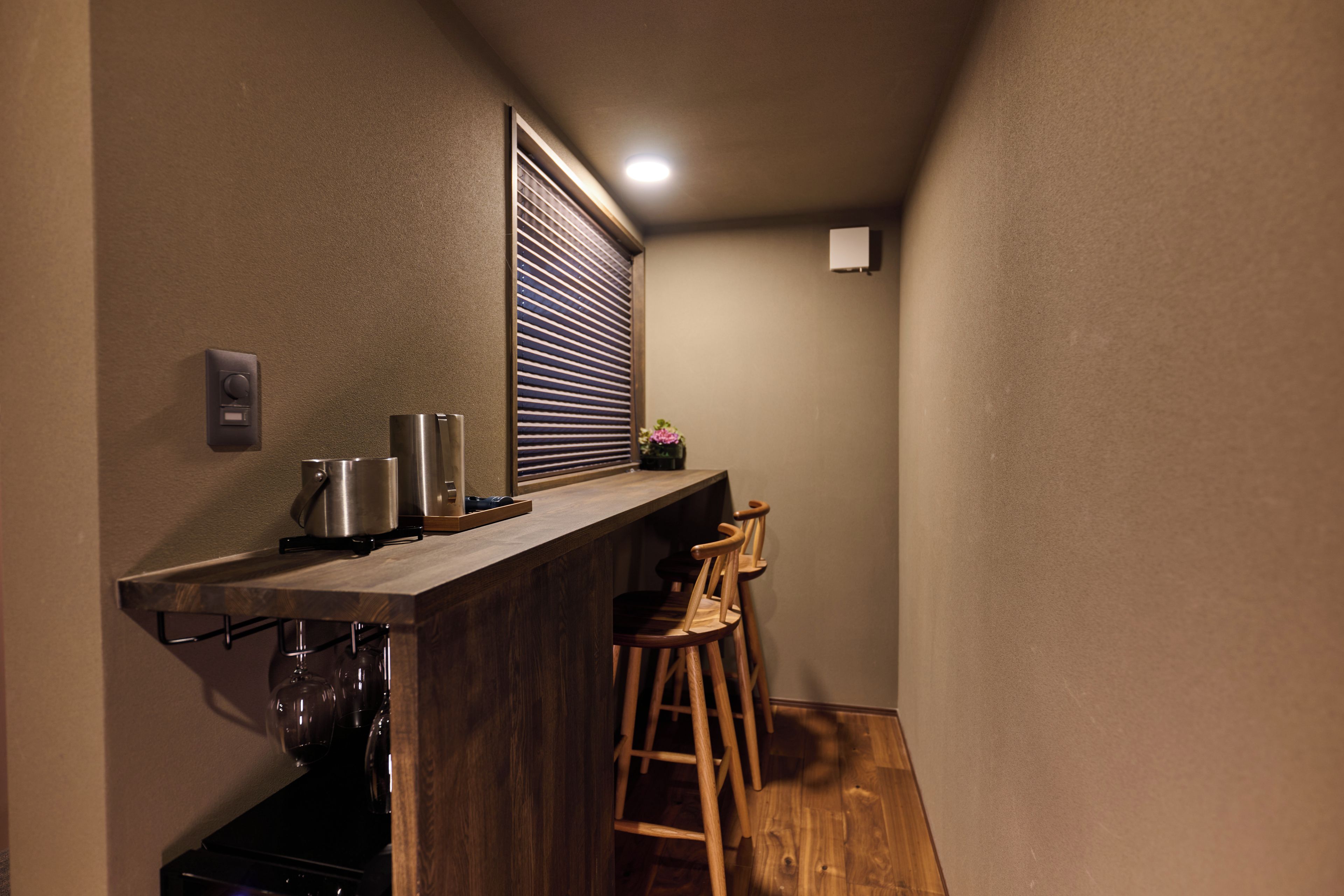 A narrow kitchen with a wooden countertop, a window with blinds, a small sink, and two wooden stools.