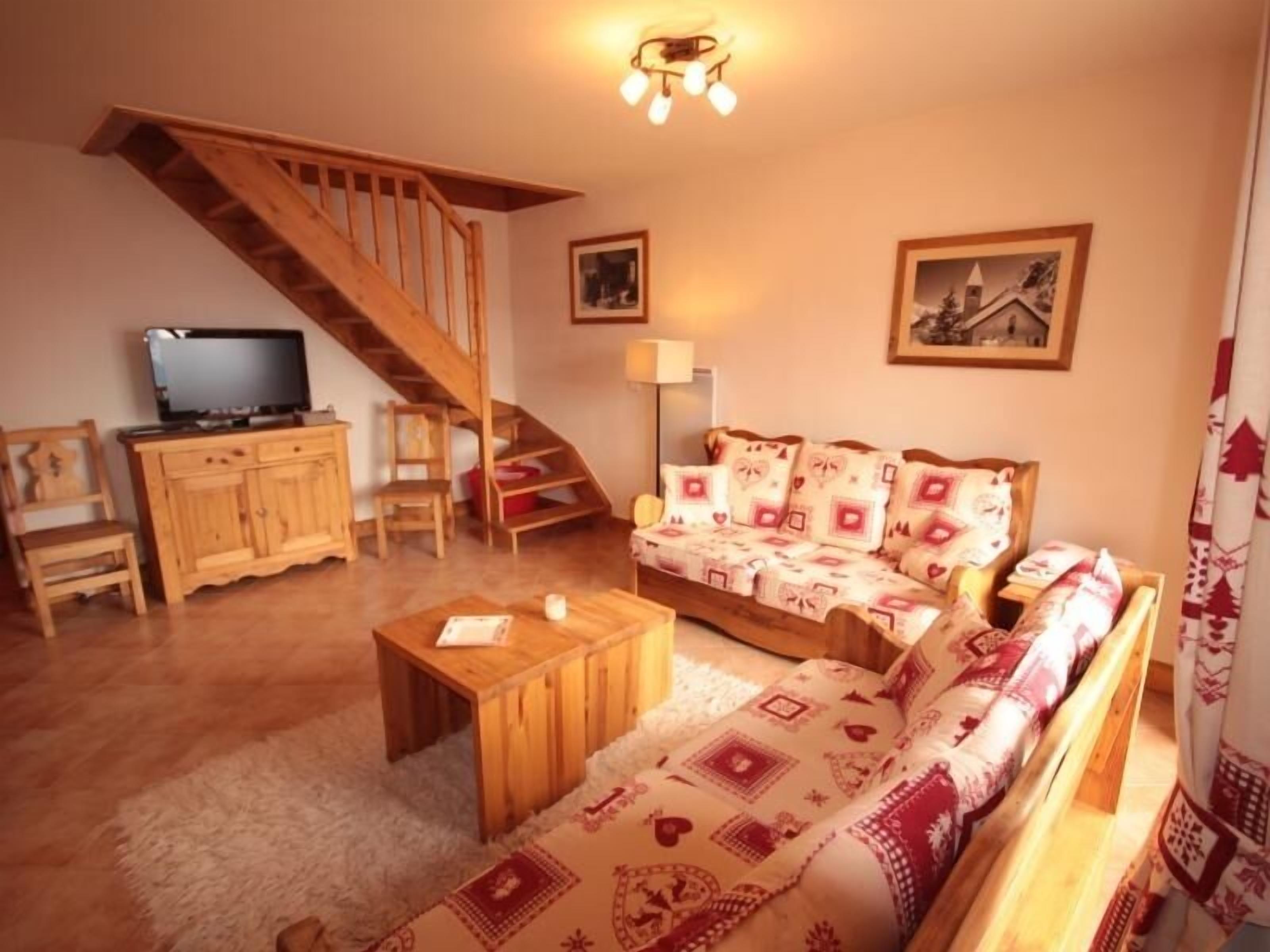 Uma sala de estar aconchegante com uma escada de madeira, um sofá com almofadas estampadas, uma televisão sobre um aparador de madeira e uma pequena mesa com uma vela.