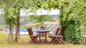 An outdoor patio with a wooden table and chairs, surrounded by greenery and a view of a swimming pool. - Villa Bacco (Peccioli)