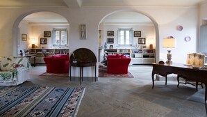 A spacious living room with a fireplace, red armchairs, a wooden coffee table, and a dining table with chairs. There are bookshelves filled with books and decorative items, and a large window providing natural light. - Villa Bacco (Peccioli)