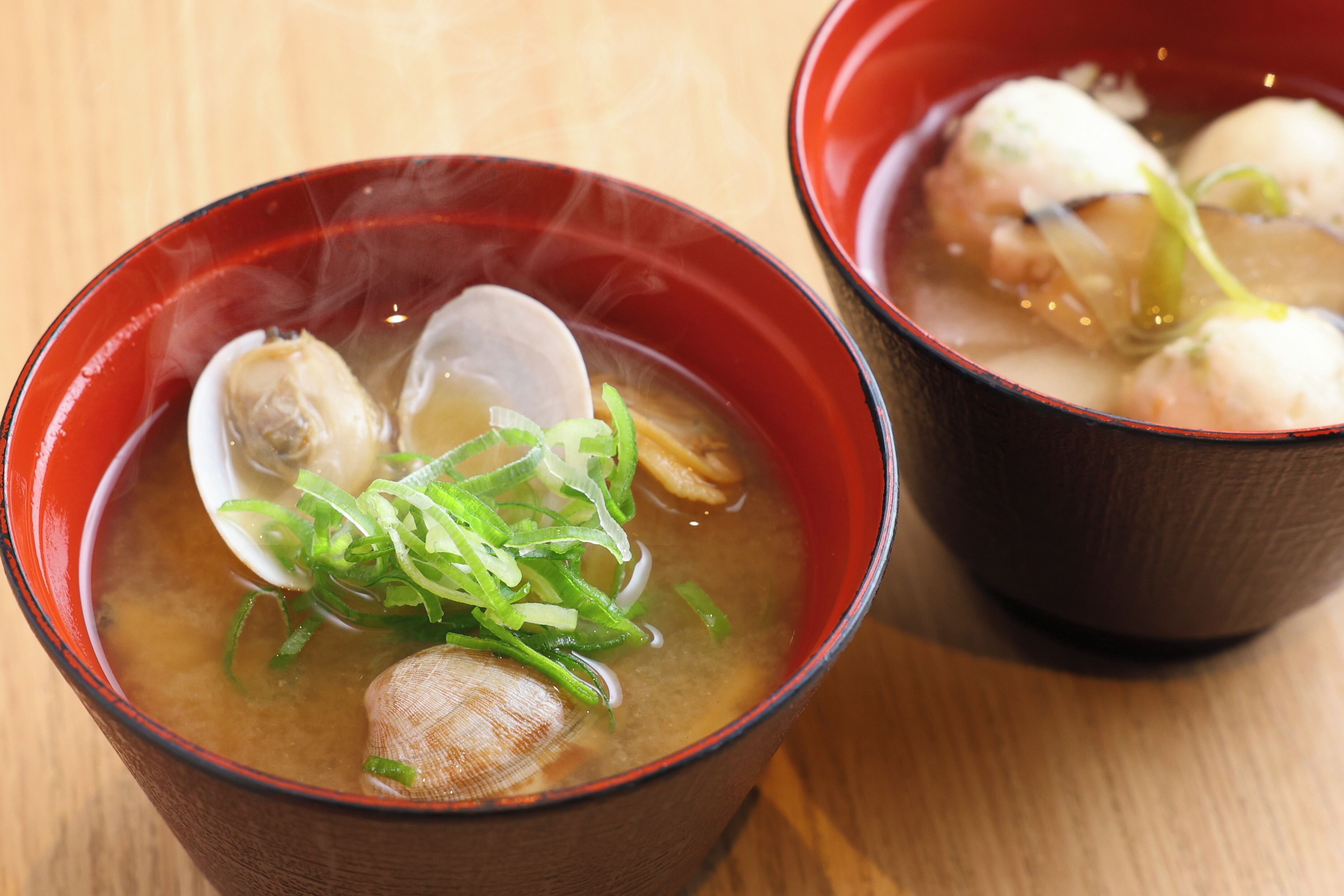 Two bowls of soup with clams and green onions, served in red bowls on a wooden table.