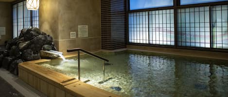 Un área de aguas termales con una terraza de madera, un muro de piedra y una vista a través de una ventana con diseño de cuadrícula.