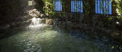 Un área de aguas termales con un muro de piedra, plantas de bambú y una cascada.