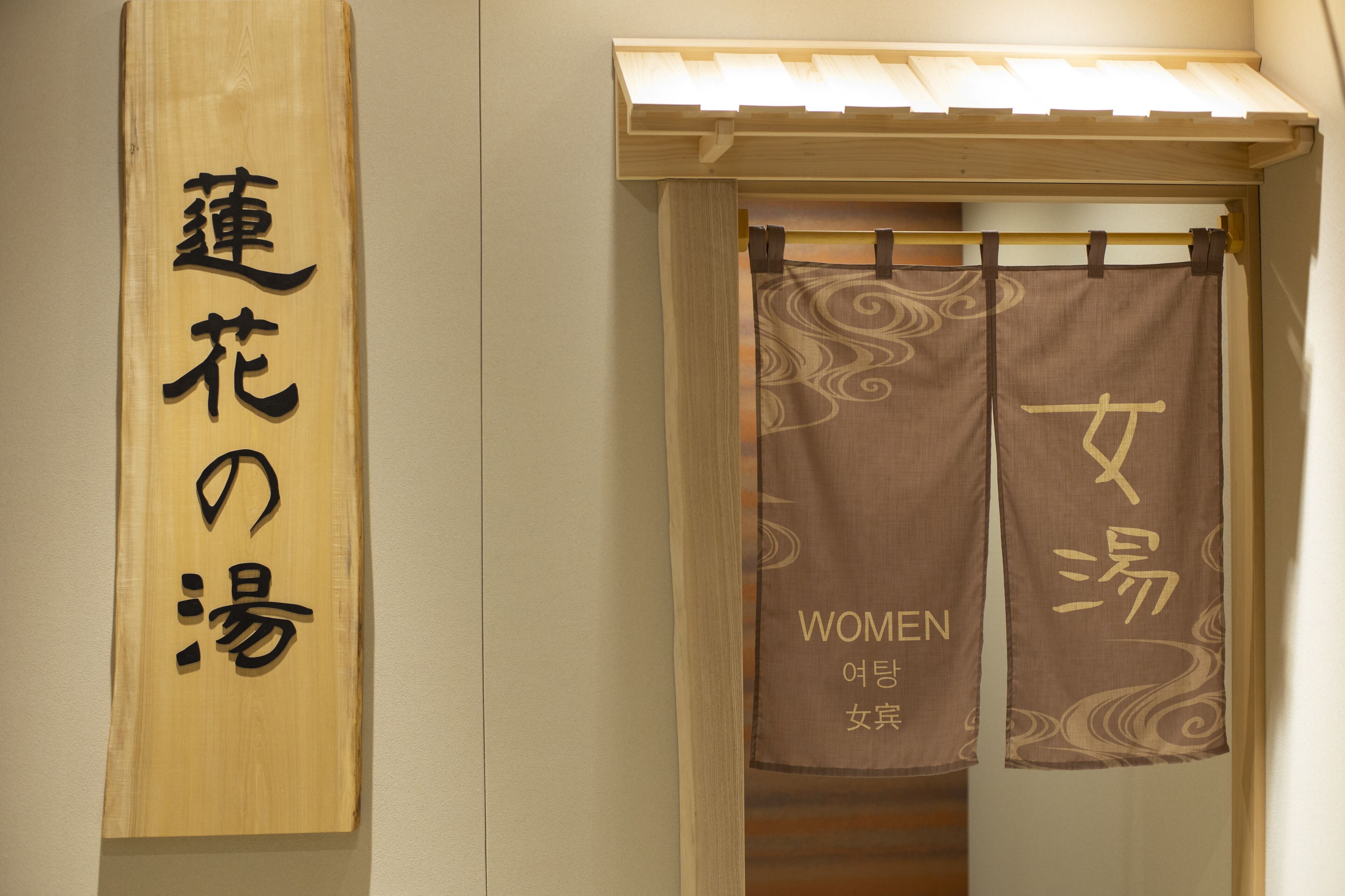 A wooden sign with Japanese characters, a door with a curtain featuring the word "WOMEN" and additional text, and a wooden structure above the door.