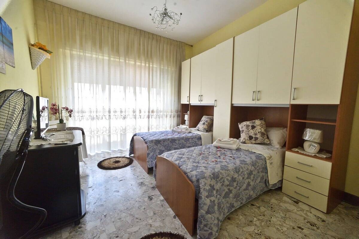 A hotel room with two beds, a desk, a chair, a fan, and a wardrobe.