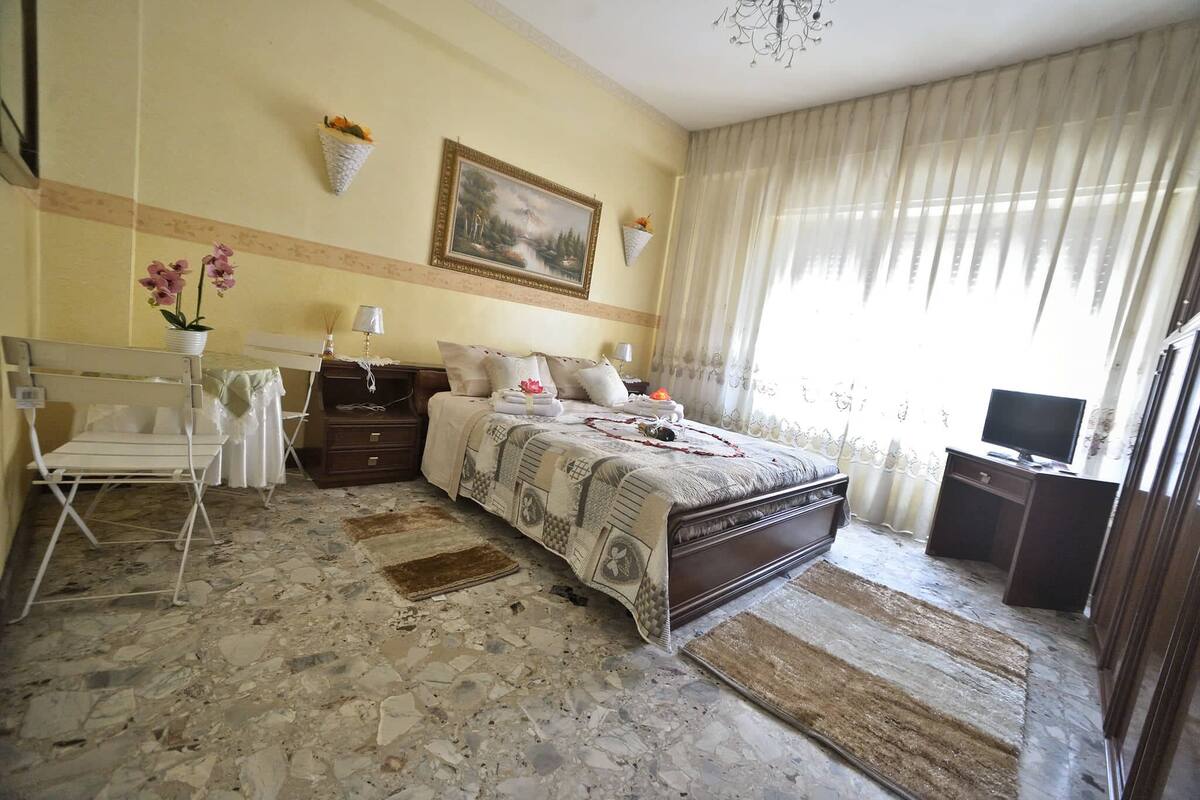 A bedroom with a bed, bedside tables, a desk, a chair, a TV, and a painting on the wall.
