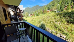 Ein Balkon mit Korbmöbeln und einem kleinen Tisch, der auf eine gebirgige Landschaft mit Häusern und Grünblicken blickt.