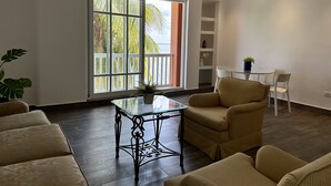 Una sala de estar con una mesa de centro de vidrio, sillones color beige, una mesa de comedor blanca con sillas y vistas a palmeras a través de amplios ventanales.