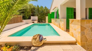 Une piscine rectangulaire à l’eau d’un bleu limpide, entourée d’un mur de pierre et de volets verts, avec un chapeau de paille et un livre posés sur le bord.