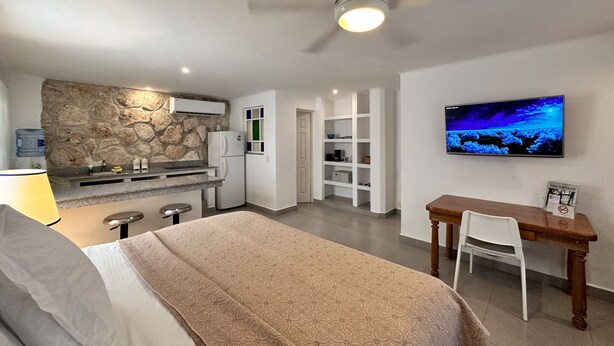 A bedroom with a bed, a desk, a chair, a TV, a ceiling fan, and a kitchen area with a refrigerator and a stone wall. - COZY STUDIO WITH FULL KITCHEN ★ AMAZING LOCATION (Cancún)