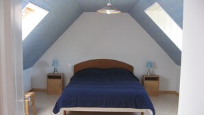 Un dormitorio con techo inclinado, una cama con colcha azul, dos mesitas de noche con lámparas y un tragaluz.