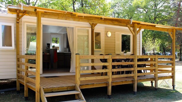Een houten terras met een helling die leidt naar een overdekte zithoek, een kleine keuken is zichtbaar en er is uitzicht op bomen in de verte.