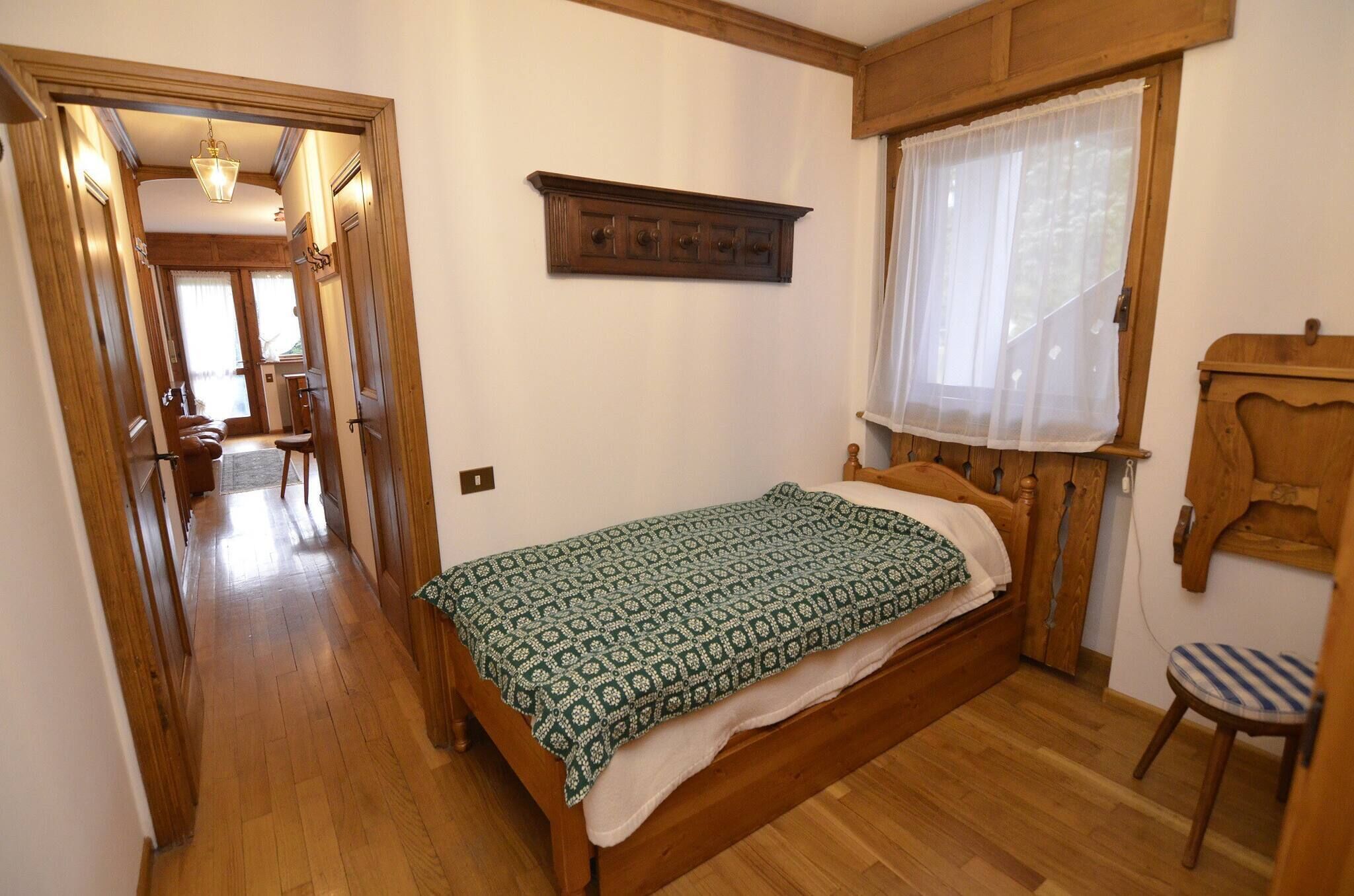 A bedroom with a wooden bed, a small chair, a wooden shelf, and a window with a sheer curtain.