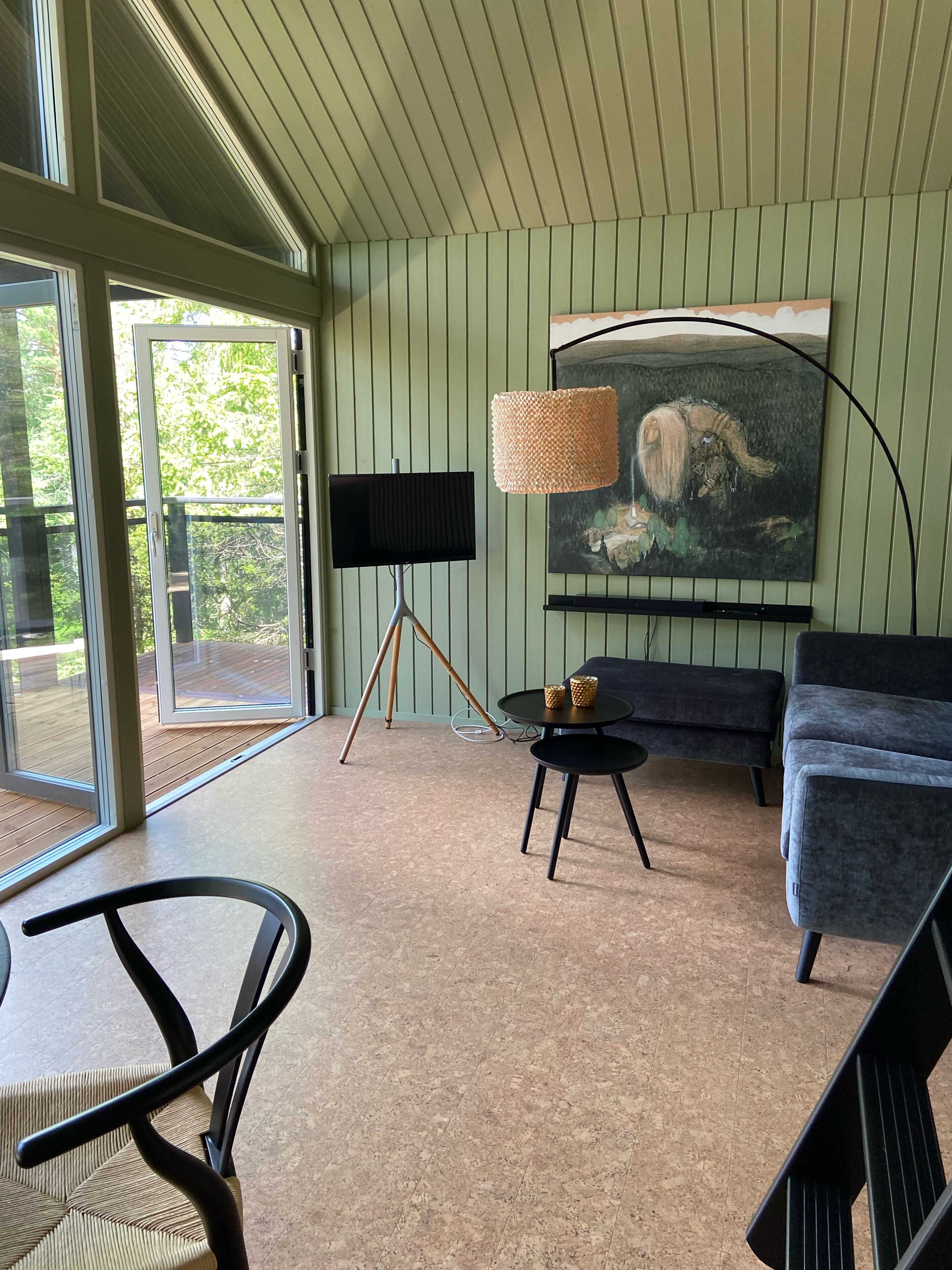 A modern living room with a large window, a black TV on a tripod stand, a round table with two candles, a dark blue sofa, and a painting of a landscape.