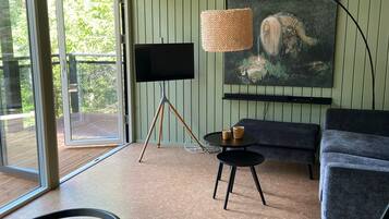 A modern living room with a large window, a black TV on a tripod stand, a round table with two candles, a dark blue sofa, and a painting of a landscape.