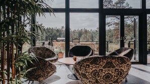 A modern outdoor seating area with patterned chairs and a small table, surrounded by large windows offering a view of trees and a distant building. - Mavens Kustnära Stugor (Lärbro)