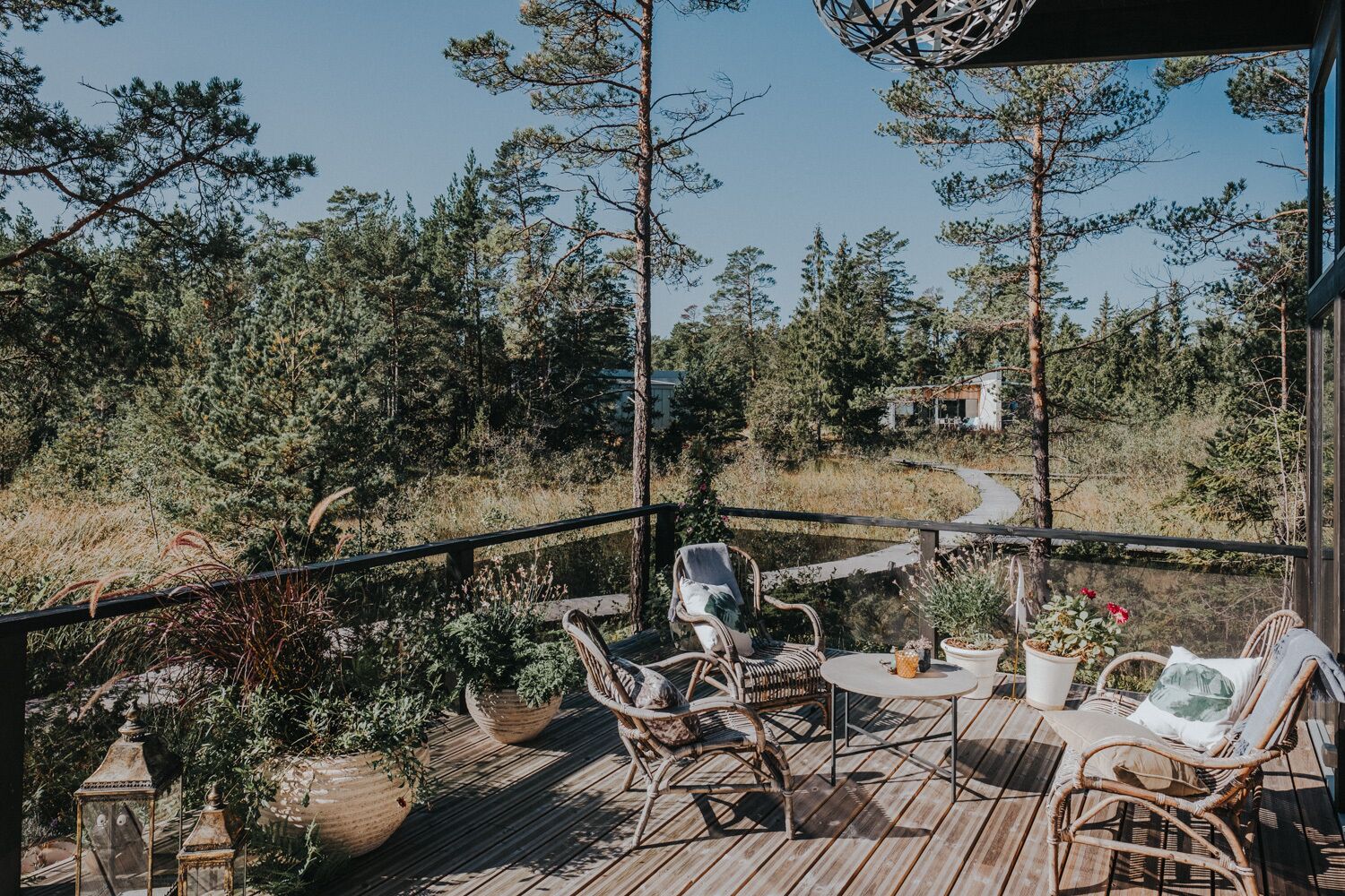 A wooden deck with wicker chairs and a small table, surrounded by potted plants and overlooking a forested area.