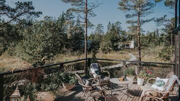 A wooden deck with wicker chairs and a small table, surrounded by potted plants and overlooking a forested area.