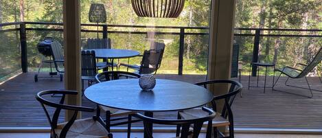 Una amplia terraza al aire libre con una mesa redonda y sillas, rodeada de grandes ventanales que dan a una zona boscosa.