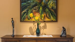 Una pintura de una escena tropical con un jarrón de flores amarillas sobre una mesa de madera.