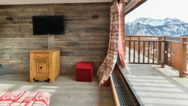 A room with a wooden wall, a television mounted on it, a wooden cabinet, a red stool, and a balcony with a view of snow-capped mountains. - Renovated 2BR apartment at ski slopes with parking - Courchevel 1850 (Courchevel 1850)