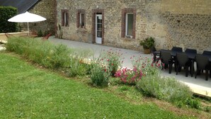 A stone building with a door and windows, surrounded by a garden with flowers and chairs.