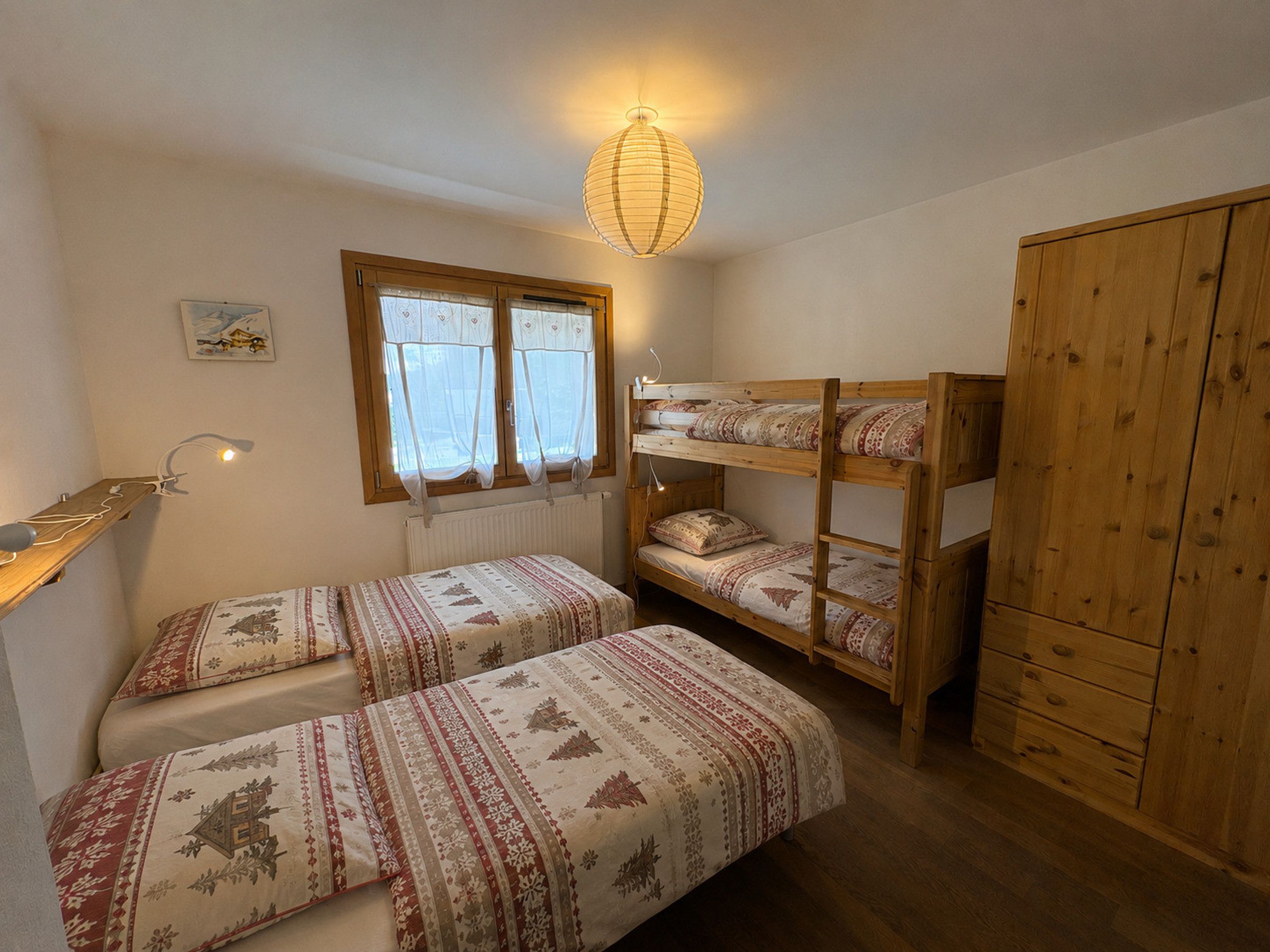 A bedroom with two single beds, a bunk bed, a wooden wardrobe, a radiator, a window with curtains, and a hanging light fixture.
