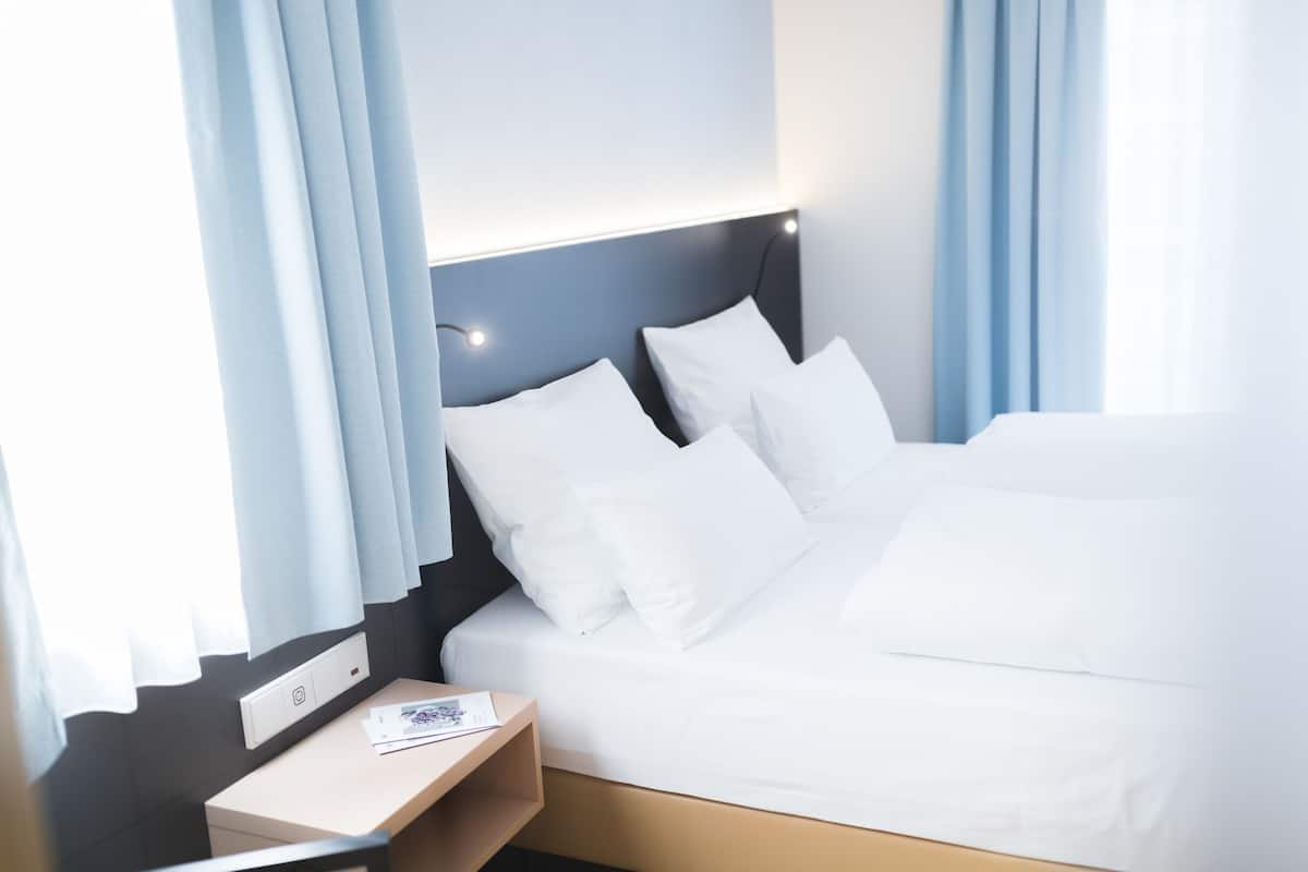 A hotel room with a bed, a bedside table, and a window with curtains.