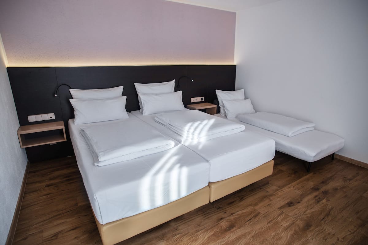 A modern hotel room with a large bed, a smaller bed, and a daybed. There are bedside tables and a wall-mounted lamp.