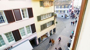 En brostensbelagt gade med fodgængere, bygninger med smalle vinduesrammer, og en butik med et skilt.
