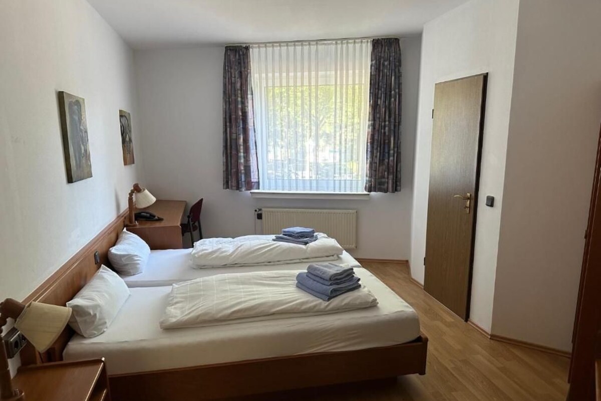 A hotel room with two single beds, a desk with a chair, a radiator, and a window with curtains.
