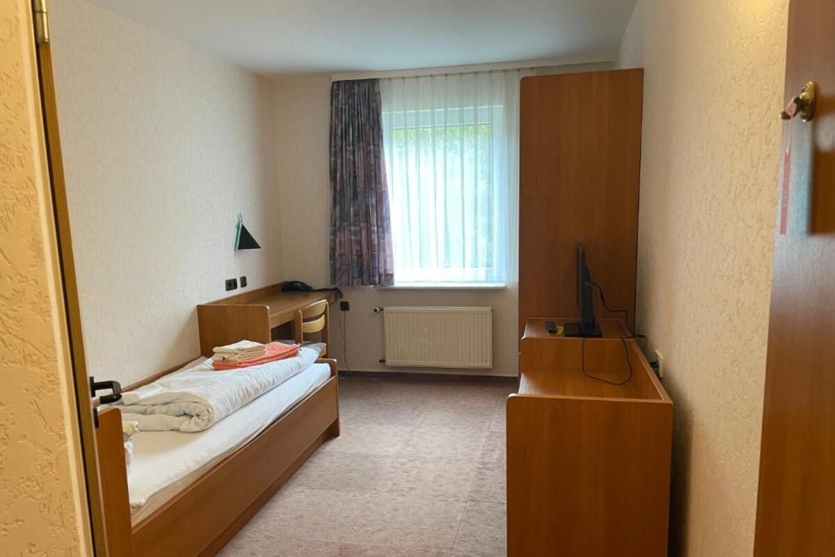 A small, compact room with a single bed, a wooden desk, a radiator, and a window with curtains.