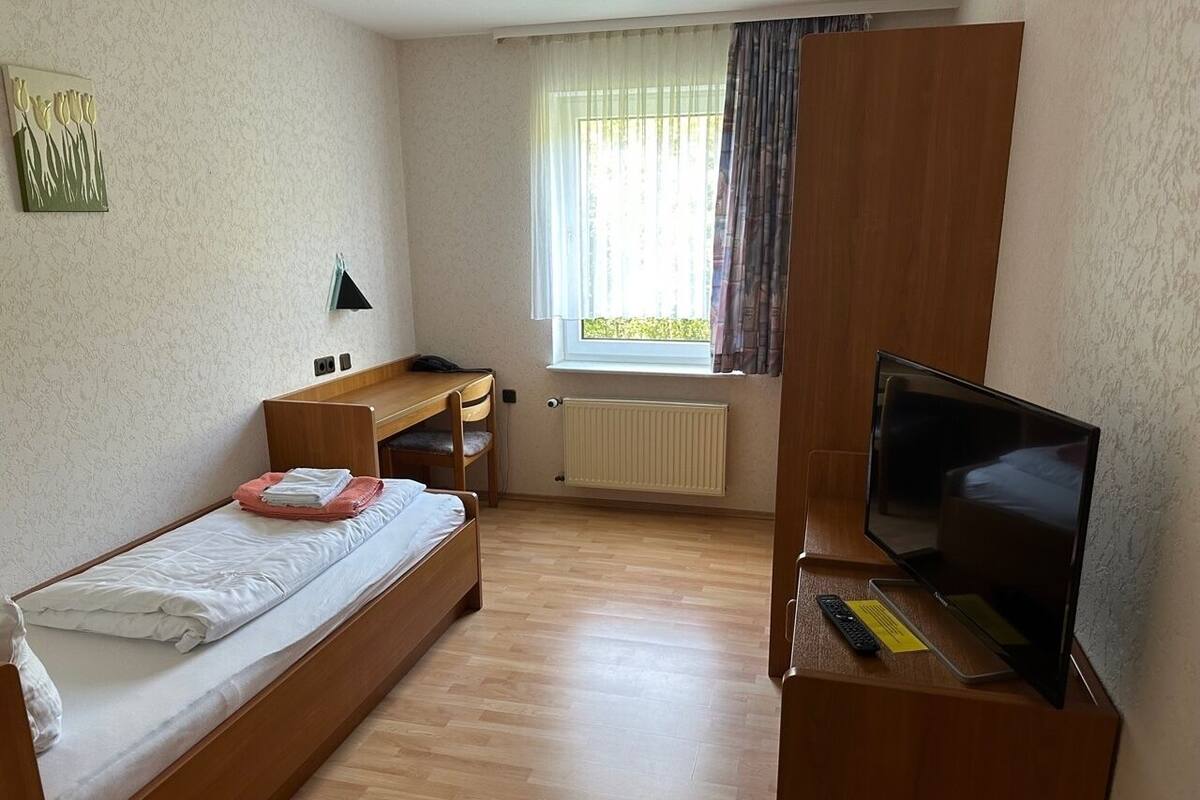 A small, tidy room with a single bed, a desk, a chair, a television, and a window with curtains.
