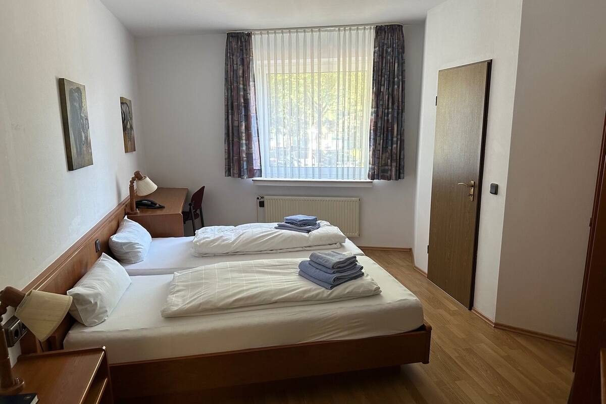 A hotel room with two single beds, a desk, a chair, a radiator, and a window with curtains.