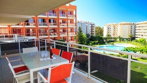 A balcony with a glass table and chairs, overlooking a swimming pool and surrounding greenery.