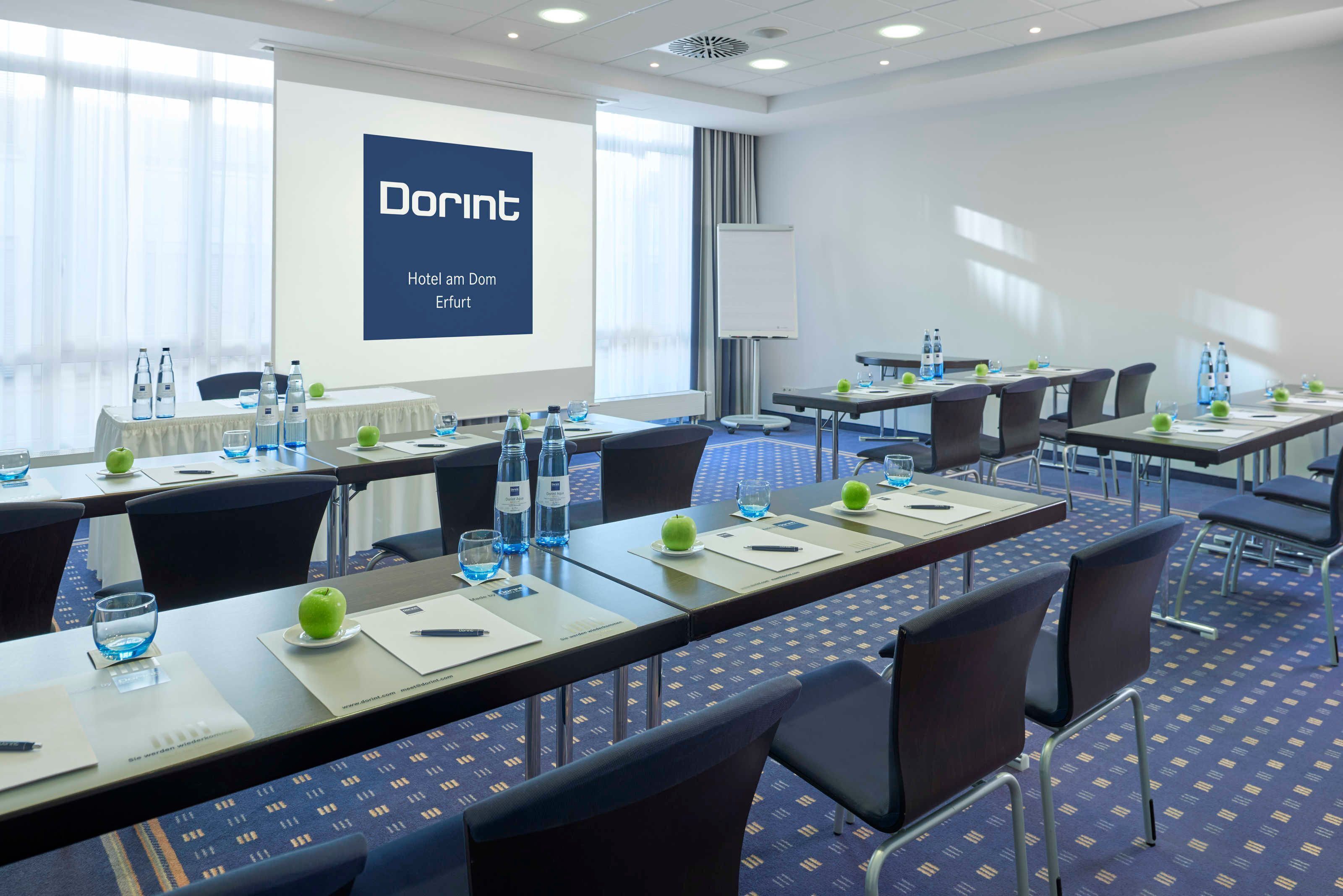 Een vergaderruimte met tafels klaar voor vergaderingen, een projectorscherm waarop 'Domint Hotel am Dom Erfurt' staat en een whiteboard.