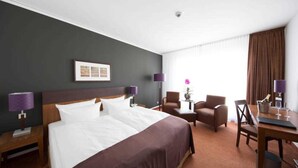 Une chambre d’hôtel avec un grand lit, deux fauteuils, une petite table ornée d’un vase de fleurs et un bureau avec une lampe et une bouteille de champagne.