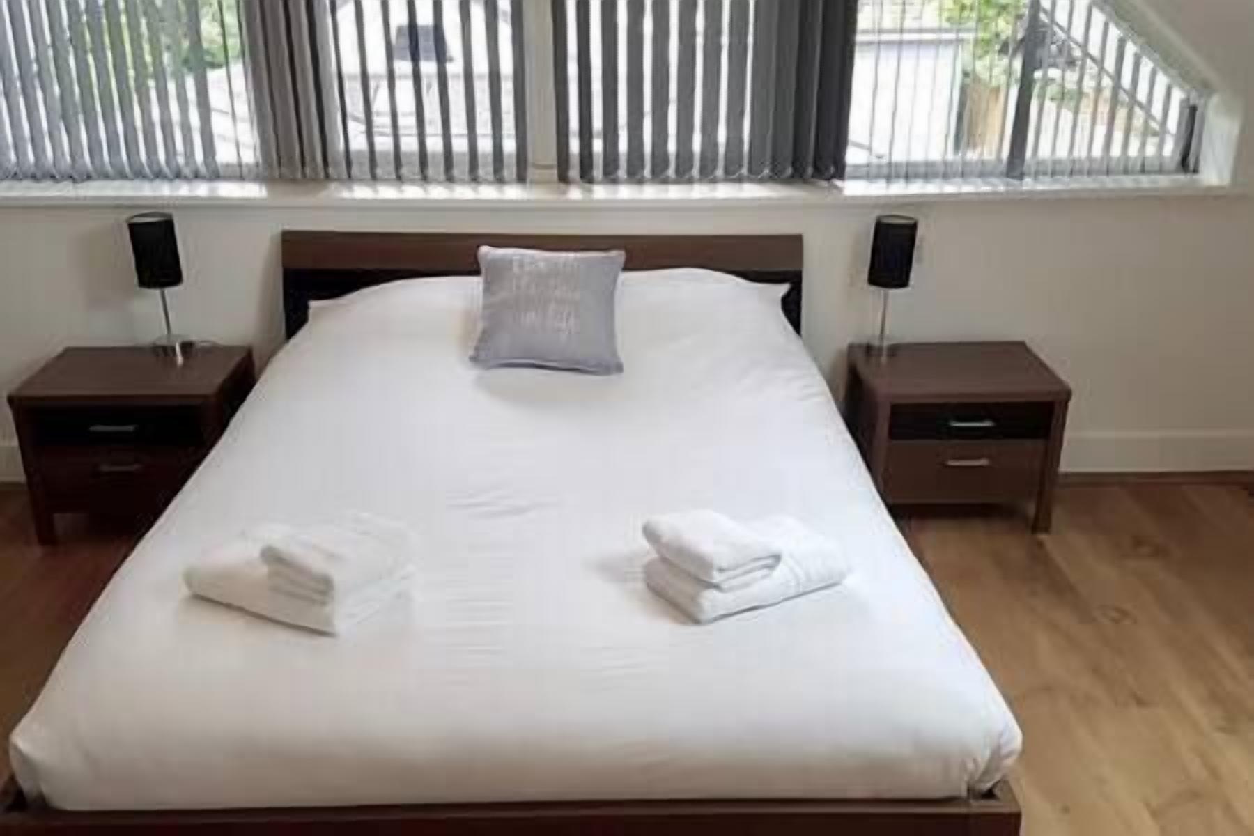 A neatly made bed with white linens and two pillows, flanked by two bedside tables with lamps, in a room with wooden flooring and a window with blinds.